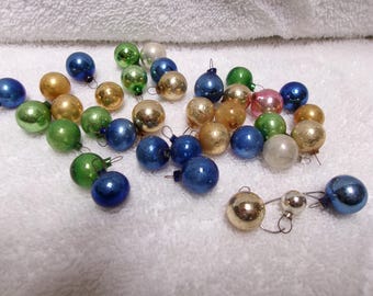 Pode incluir: Uma coleção de pequenos enfeites de Natal redondos em várias cores, incluindo ouro metálico, azul brilhante, verde vibrante, prata e rosa. Cada enfeite esférico possui uma pequena tampa de metal no topo para pendurar, com um acabamento brilhante.