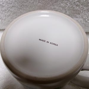 Op de afbeelding: Een witte keramische mok met een lichtgrijze rand en oor. De onderkant van de mok heeft de tekst "MADE IN KOREA" in zwarte letters. De mok staat op een wit, getextureerd oppervlak.