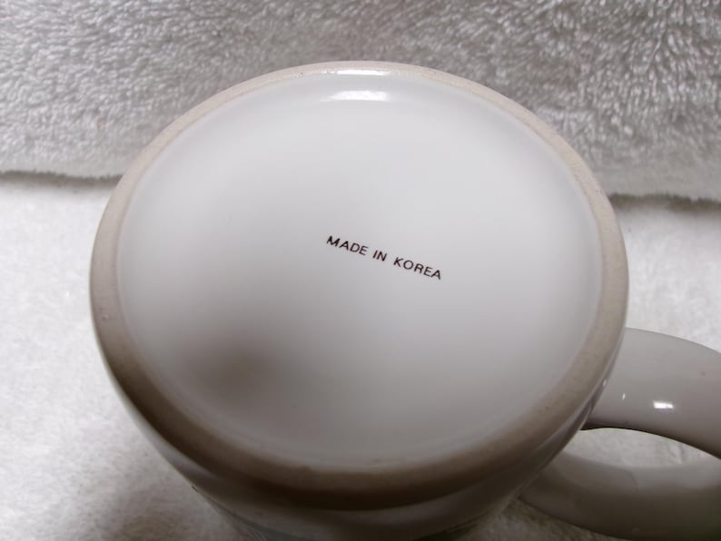 Op de afbeelding: Een witte keramische mok met een lichtgrijze rand en oor. De onderkant van de mok heeft de tekst "MADE IN KOREA" in zwarte letters. De mok staat op een wit, getextureerd oppervlak.