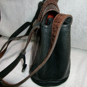 May include: A black leather shoulder bag with brown accents. The bag has a textured surface and a long, adjustable strap. The top flap and strap feature a brown, patterned design. The interior is a bright orange color.