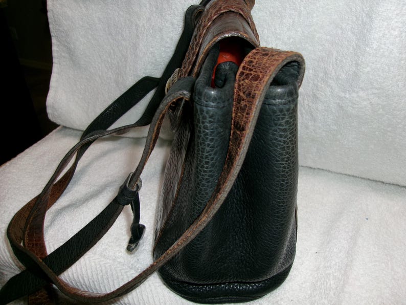 May include: A black leather shoulder bag with brown accents. The bag has a textured surface and a long, adjustable strap. The top flap and strap feature a brown, patterned design. The interior is a bright orange color.