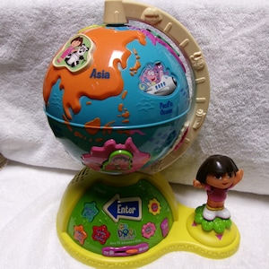 May include: A yellow and blue Dora the Explorer interactive globe toy. The globe features illustrations of Dora and other characters, with the continents labeled. A Dora figurine stands on the base. The base has buttons and the word "Enter".