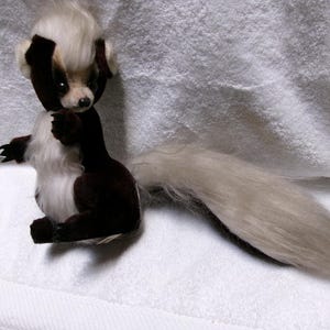 May include: A handmade plush skunk toy with a black and white color scheme. The skunk has a fluffy white tail, a white stripe down its back, and a white patch on its head. The toy is sitting on a white surface.