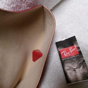 Op de afbeelding: Een open rode brillenkoker met een beige interieur en een rode driehoekige sluiting. Een zwart-wit Ray-Ban-label staat naast de koker. Het label heeft het Ray-Ban-logo en de tekst "GENUINE SINCE 1937".