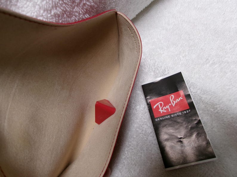 Op de afbeelding: Een open rode brillenkoker met een beige interieur en een rode driehoekige sluiting. Een zwart-wit Ray-Ban-label staat naast de koker. Het label heeft het Ray-Ban-logo en de tekst "GENUINE SINCE 1937".