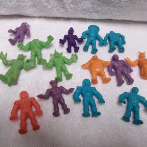 May include: A collection of colorful, small monster figurines in various poses. The figures are in shades of green, purple, orange, red, and blue. The toys are made of plastic and are on a white surface.