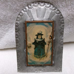 May include: A rectangular wall hanging featuring a central print of a person in a dark hat and teal dress holding a basket and staff, set against a blue and white cloudy background. The print is framed in wood and mounted on a silver metal backing.