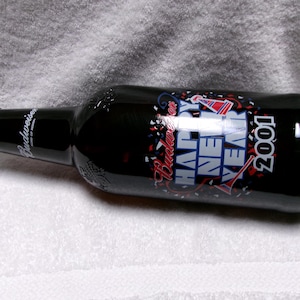 May include: A black Budweiser beer bottle with a "Happy New Year 2001" label. The label features red, white, and blue accents with confetti and a party hat design. The bottle is lying on a white surface.