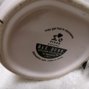 May include: The underside of a white ceramic mug featuring black printed text that reads: may get hot in microwave, Pixar, Disney/Pixar, Rae Dunn, Artisan Collection by Magenta, -181-, dishwasher safe, made in china. A black ribbon is attached to the side.