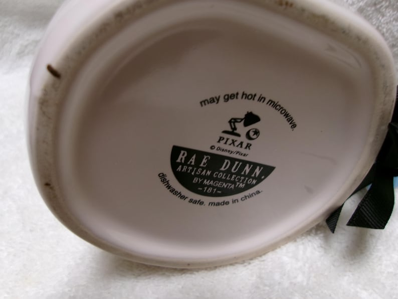 May include: The underside of a white ceramic mug featuring black printed text that reads: may get hot in microwave, Pixar, Disney/Pixar, Rae Dunn, Artisan Collection by Magenta, -181-, dishwasher safe, made in china. A black ribbon is attached to the side.