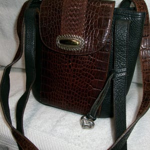 May include: A brown and black crossbody bag with a textured, crocodile-like pattern. The bag features a flap closure with a silver-toned metal accent and a heart-shaped charm. The adjustable strap is a combination of brown and black.