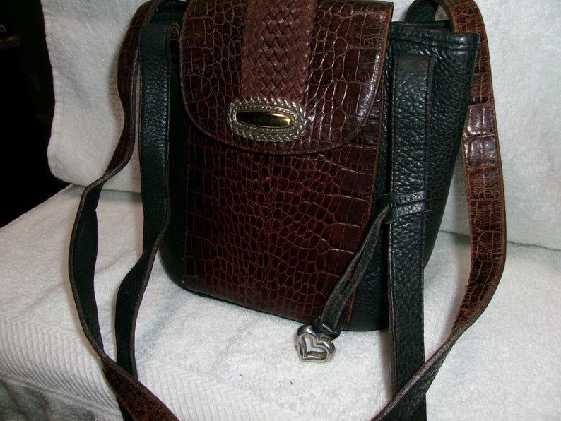 May include: A brown and black crossbody bag with a textured, crocodile-like pattern. The bag features a flap closure with a silver-toned metal accent and a heart-shaped charm. The adjustable strap is a combination of brown and black.