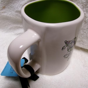May include: A white ceramic mug with a bright green interior, featuring a graphic of a space ranger character on the side. A blue rectangular tag is attached to the handle with a black ribbon, and the mug has a rounded square shape.