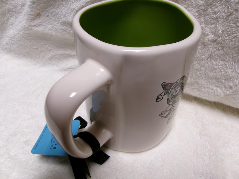 May include: A white ceramic mug with a bright green interior, featuring a graphic of a space ranger character on the side. A blue rectangular tag is attached to the handle with a black ribbon, and the mug has a rounded square shape.