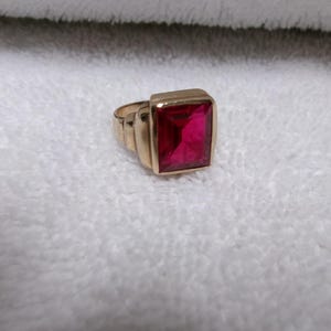 May include: A gold-toned ring featuring a large, rectangular, faceted deep pink gemstone set in a smooth metal bezel. The band has a stepped, geometric design on the sides, leading up to the prominent, vibrant pink stone at the center.