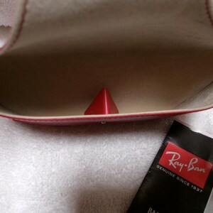 Op de afbeelding: Open rode brillenkoker met een rood piramidevormig object erin. De binnenkant is beige. Een zwart Ray-Ban-label met rode en witte tekst staat rechtsonder. De koker ligt op een wit, getextureerd oppervlak.