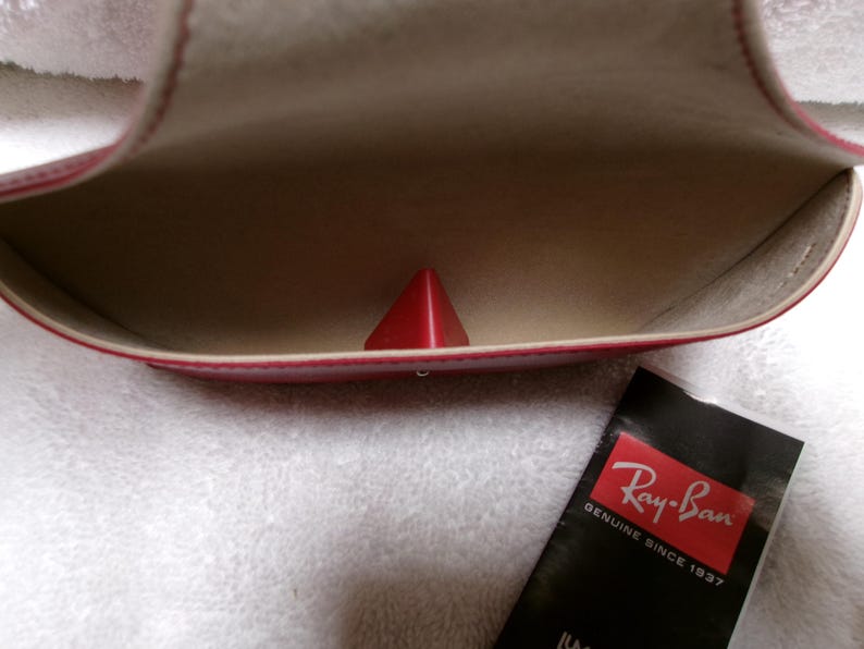 Op de afbeelding: Open rode brillenkoker met een rood piramidevormig object erin. De binnenkant is beige. Een zwart Ray-Ban-label met rode en witte tekst staat rechtsonder. De koker ligt op een wit, getextureerd oppervlak.