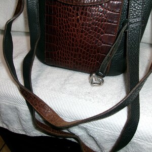 May include: A brown and black leather crossbody bag with a textured, crocodile-like pattern on the front flap. The bag features a silver heart-shaped charm and a long, adjustable strap.