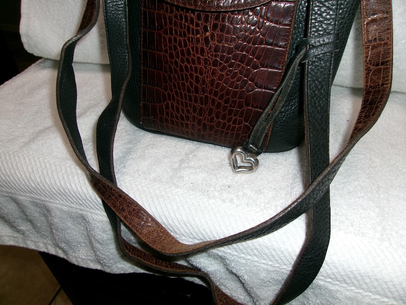 May include: A brown and black leather crossbody bag with a textured, crocodile-like pattern on the front flap. The bag features a silver heart-shaped charm and a long, adjustable strap.