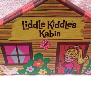 May include: A Liddle Kiddles Kabin toy house featuring a log cabin design with a red and brown roof. The front displays the text Liddle Kiddles Kabin, a pink butterfly, a white rabbit, and a cartoon illustration of a blonde character by a door.