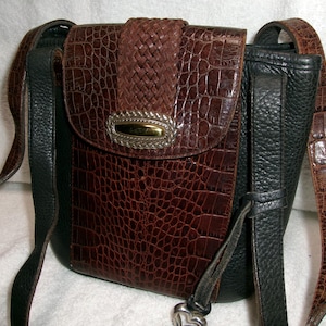 May include: A brown and black crossbody bag with a faux crocodile skin pattern. The bag features a woven flap, a silver and gold-toned clasp, and adjustable straps. The bag's dimensions are approximately 8 inches in height and 7 inches in width.