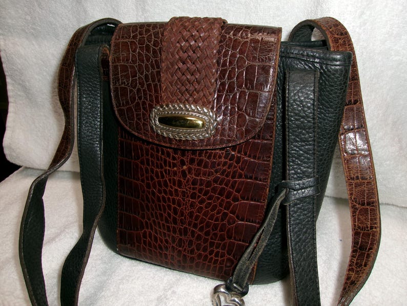May include: A brown and black crossbody bag with a faux crocodile skin pattern. The bag features a woven flap, a silver and gold-toned clasp, and adjustable straps. The bag's dimensions are approximately 8 inches in height and 7 inches in width.