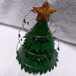 May include: A miniature Christmas tree figurine. The green tree is adorned with a gold star on top and a string of colorful lights. The tree is made of a textured material.