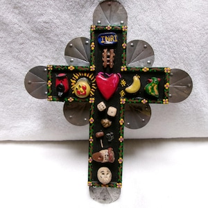 May include: A black wooden cross decorated with small colorful figurines including a red heart, a yellow sun, a yellow moon, dice, and a blue plaque with the text INRI. The cross features metal circular accents at the ends and edges.