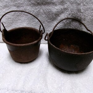 May include: Two small, antique, cast iron pots with arched handles. The pots are dark brown with a rustic appearance. The pot on the left is cylindrical, while the pot on the right is more rounded. Both pots are empty.