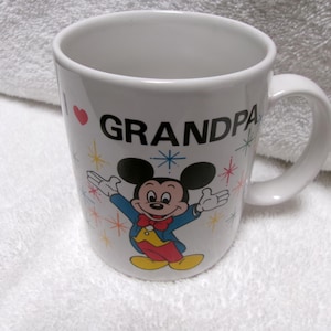 Op de afbeelding: Witte keramische mok met een gebogen handvat. De mok heeft de tekst "I ❤️ GRANDPA" met een rood hart en een kleurrijke illustratie van Mickey Mouse. Het ontwerp bevat sterretjes.