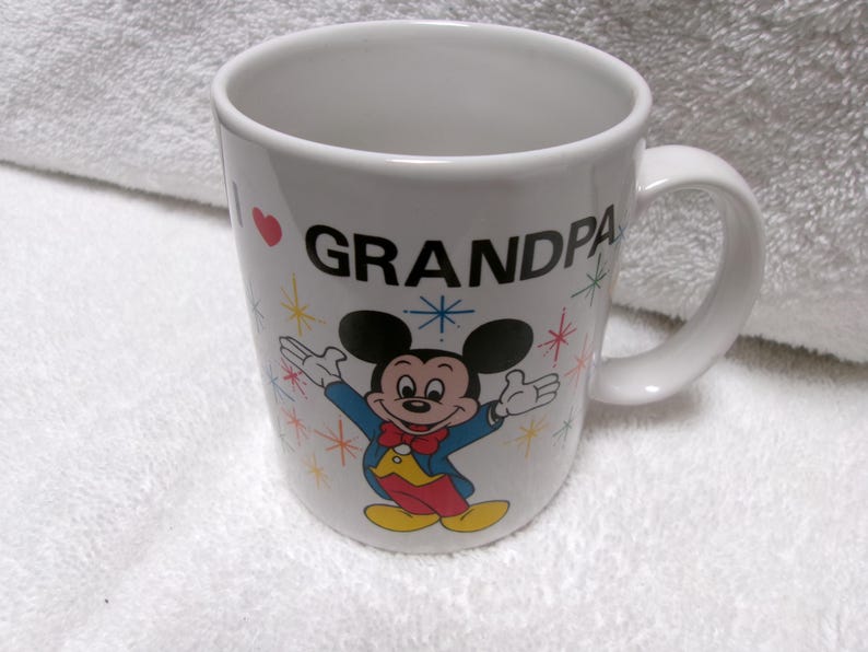 Op de afbeelding: Witte keramische mok met een gebogen handvat. De mok heeft de tekst "I ❤️ GRANDPA" met een rood hart en een kleurrijke illustratie van Mickey Mouse. Het ontwerp bevat sterretjes.