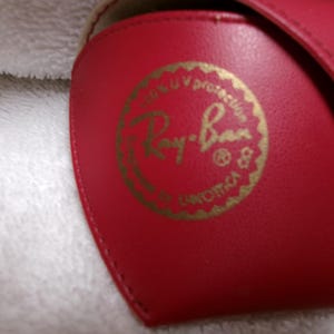 Op de afbeelding: Een rode zonnebril etui met een gouden logo. Het logo luidt "Ray-Ban" in een cirkelvormig ontwerp, met tekst die UV-bescherming aangeeft. De etui is gemaakt van een glad materiaal.