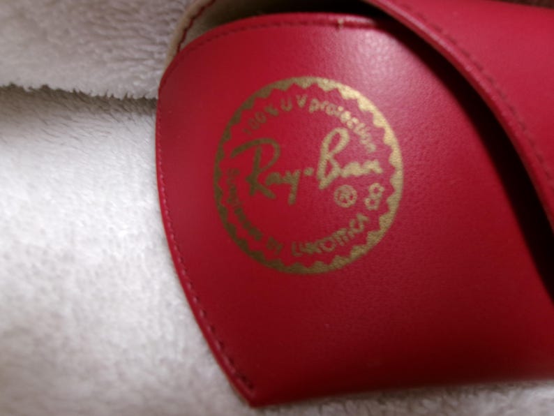 Op de afbeelding: Een rode zonnebril etui met een gouden logo. Het logo luidt "Ray-Ban" in een cirkelvormig ontwerp, met tekst die UV-bescherming aangeeft. De etui is gemaakt van een glad materiaal.