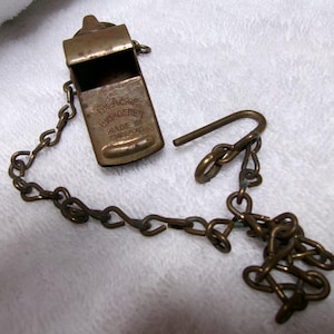 May include: Vintage brass whistle with a chain. The whistle has the text "The Acme Thunderer Made in England" on the front. The chain and whistle are a tarnished brass color. The whistle is open.