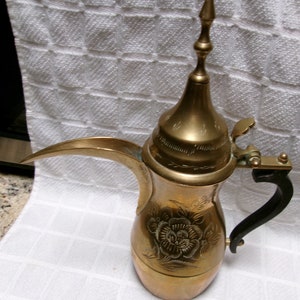 May include: A gold-colored brass coffee pot with a decorative floral design and a black handle. The pot has a tall, pointed lid with a small handle.