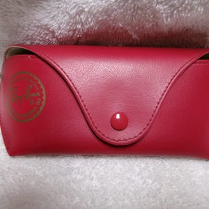 Op de afbeelding: Een rode zonnebril etui met een drukknoopsluiting. De etui heeft een gouden embleem. De etui is gemaakt van een glad materiaal en is ontworpen om brillen te beschermen.