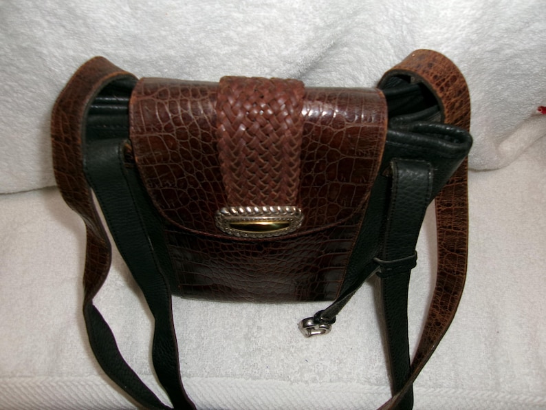 May include: A brown leather crossbody bag with a textured, crocodile-like pattern. The bag features a woven flap closure with a silver-toned clasp and a black leather strap. The bag is approximately 8 inches wide.