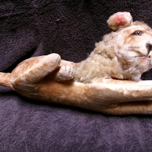 May include: A plush toy lion with a light brown and tan fur. The lion is lying on its side with its head turned to the side. The lion has a pink nose and a black nose.