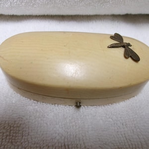 May include: An oval-shaped, cream-colored storage box with a smooth, textured surface. A small, metallic, bronze-toned dragonfly ornament is attached to the top right side of the lid. A tiny metal latch is visible on the front edge of the box.