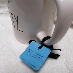 May include: A white ceramic mug with a black handle and black text that reads TY. A light blue rectangular tag is attached to the handle with a black ribbon. The tag features the text RAE DUNN. ARTISAN COLLECTION BY MAGENTA and Disney.
