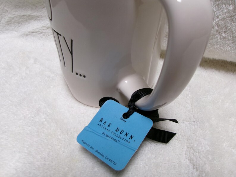 May include: A white ceramic mug with a black handle and black text that reads TY. A light blue rectangular tag is attached to the handle with a black ribbon. The tag features the text RAE DUNN. ARTISAN COLLECTION BY MAGENTA and Disney.