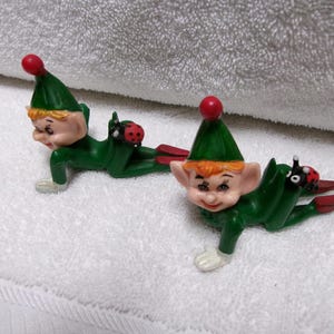 May include: Two identical elf figurines in a crawling position, each wearing a green outfit with a matching pointed hat topped with a red pom-pom. Each elf has orange hair, large ears, and a small ladybug attached to its back, with red shoes.