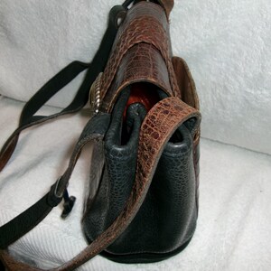 May include: A leather crossbody bag with a textured, reptile-like pattern in shades of brown and black. The bag features a long, adjustable strap and a silver-toned buckle. The interior appears to be a bright orange color.