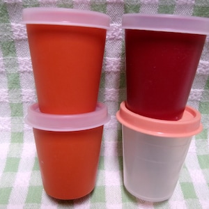 May include: Three plastic cups with lids. The cups are stacked on top of each other. The top cup is red, the middle cup is orange, and the bottom cup is white.