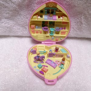 May include: A vintage Polly Pocket playset in the shape of a pink heart, open to reveal a miniature house with yellow and pink rooms. The set includes tiny furniture, accessories, and figurines. The playset is on a white surface.