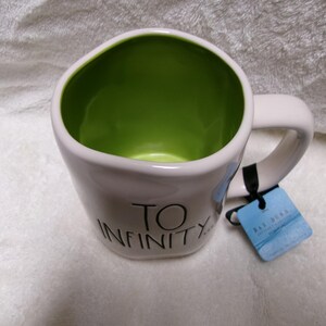 May include: A white ceramic mug with a lime green interior, featuring the black text TO INFINITY printed on the front. The mug has a rounded, irregular shape and includes a blue cardboard product tag attached to the handle with a black string.