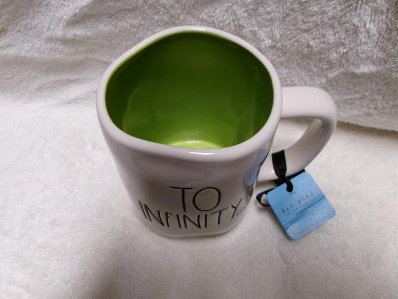 May include: A white ceramic mug with a lime green interior, featuring the black text TO INFINITY printed on the front. The mug has a rounded, irregular shape and includes a blue cardboard product tag attached to the handle with a black string.