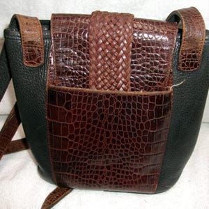 May include: A black and brown leather crossbody bag with a textured, crocodile-like pattern. The bag features a woven brown flap closure and a long, adjustable strap. The bag's design includes a front pocket and a contrasting black leather side.