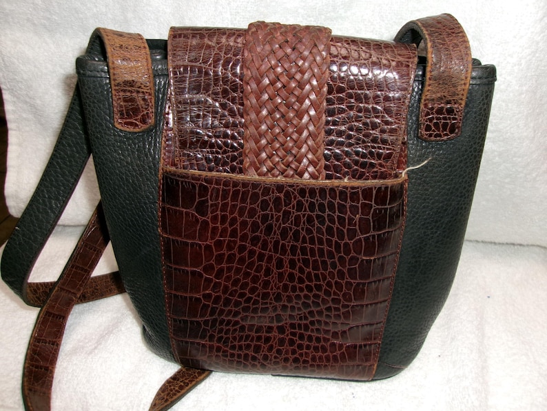May include: A black and brown leather crossbody bag with a textured, crocodile-like pattern. The bag features a woven brown flap closure and a long, adjustable strap. The bag's design includes a front pocket and a contrasting black leather side.