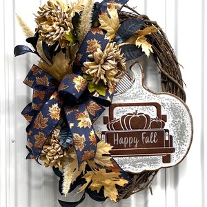 May include: A fall-themed wreath with a rustic brown grapevine base. The wreath is decorated with a blue and gold ribbon with a bow, faux fall leaves, and a white metal truck with the words "Happy Fall" on the back. The truck is filled with pumpkins.